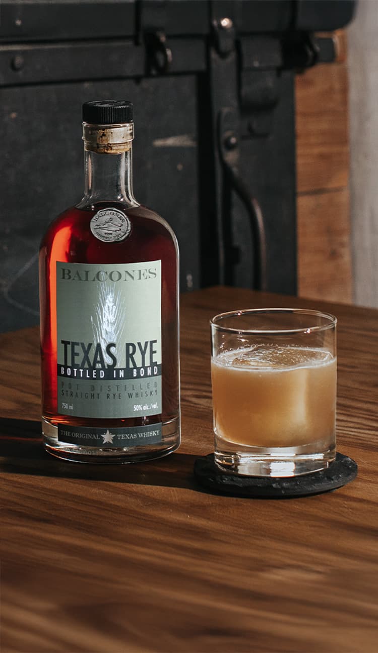 A bottle of Texas Rye Bottled in Bond next to a Banana Sour cocktail on a table.