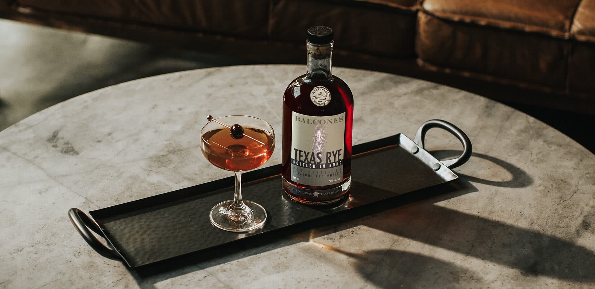 A bottle of Texas Rye Bottled in Bond next to a Black Manhattan cocktail on a table.