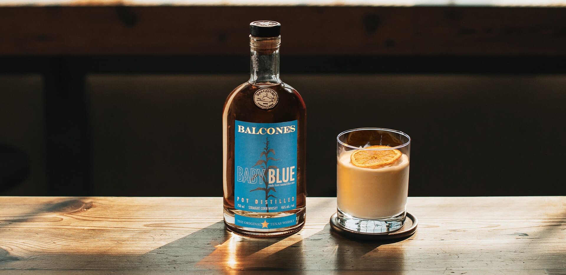 A bottle of Baby Blue Straight Corn Whisky with an It's Orange Dream, Pops cocktail on a table.