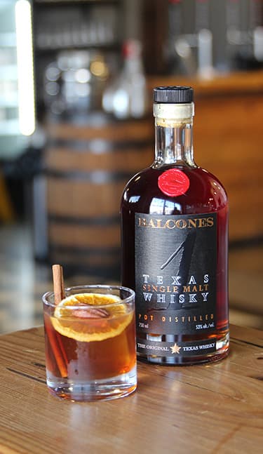 Bottle of Lineage Texas Single Malt Whisky with two glasses of Balcola cocktails on table