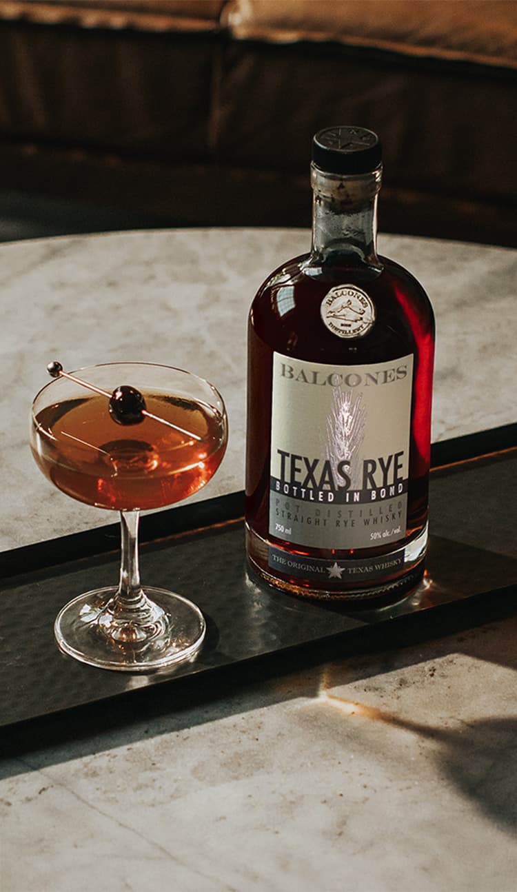A bottle of Texas Rye Bottled in Bond next to a Black Manhattan cocktail on a table.