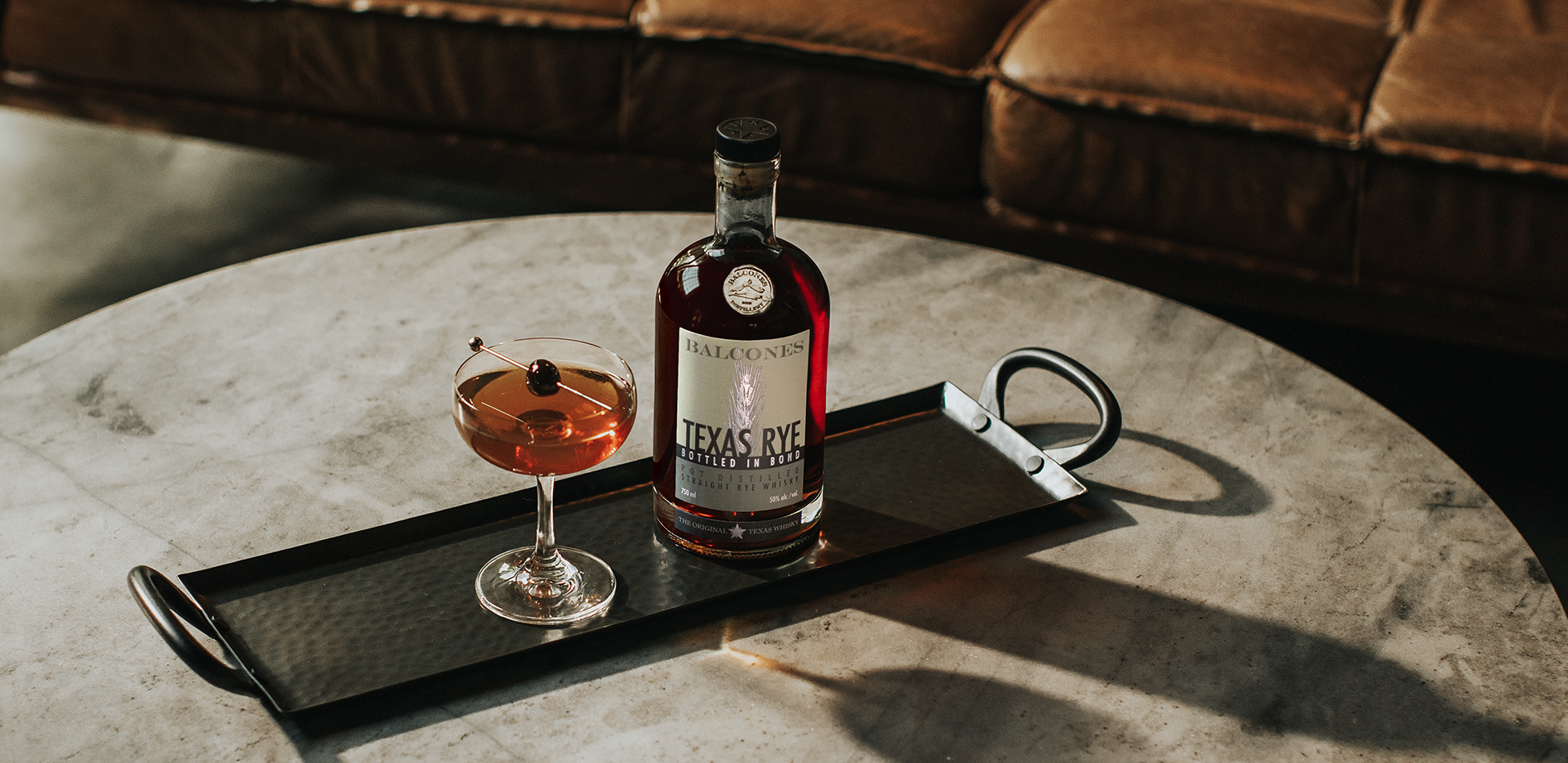 A bottle of Texas Rye Bottled in Bond next to a Black Manhattan cocktail on a table.